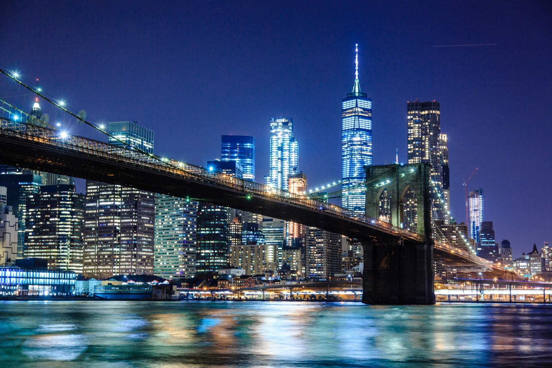 Illuminated Brooklyn Bridge with city skyline reflecting on the East River.